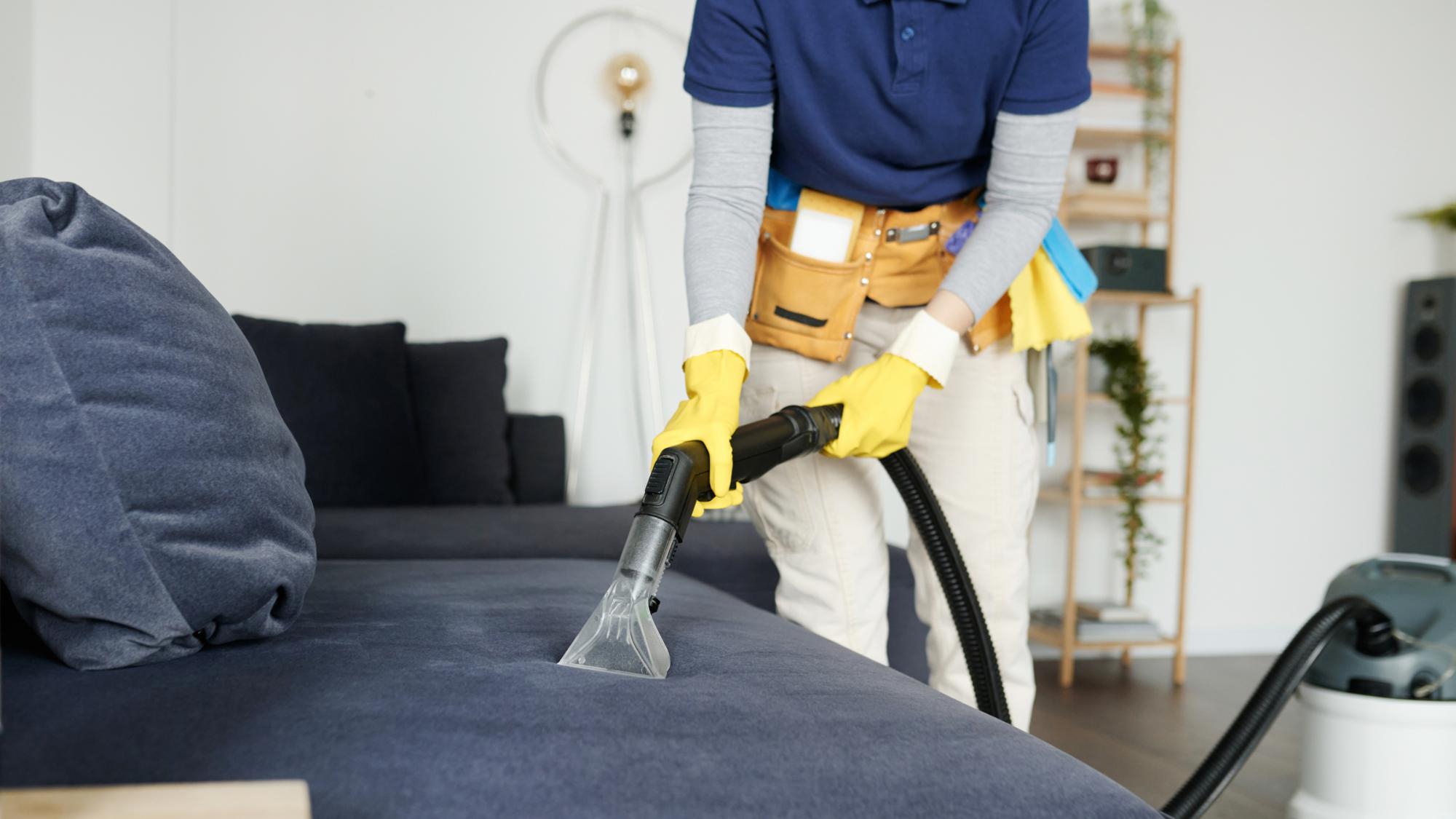 A person in a blue shirt and yellow gloves is using a vacuum cleaner attachment to clean a dark blue couch in a well-lit room with plants and furniture in the background.